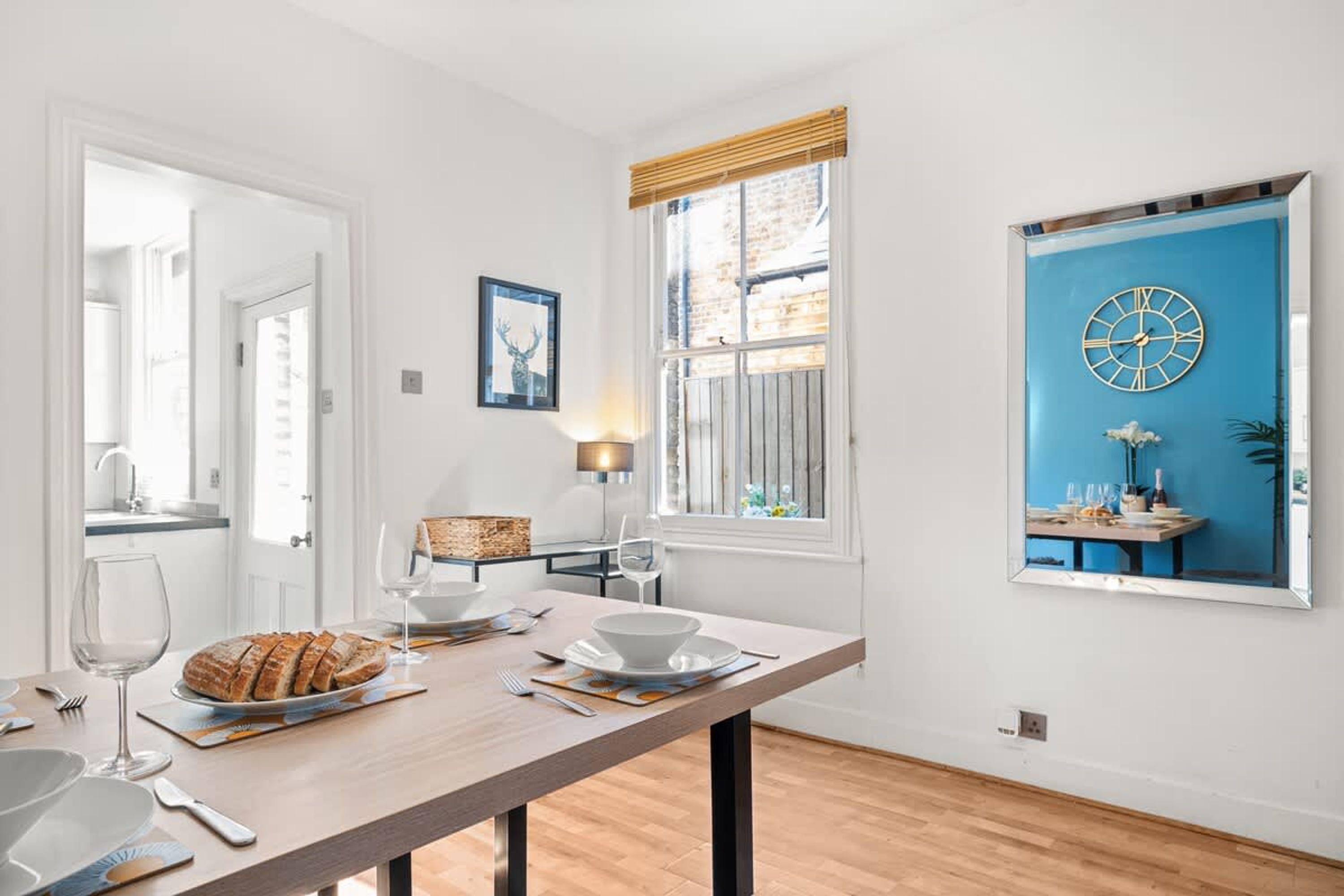 Un coin repas avec une table dressée pour deux personnes, une fenêtre donnant sur un mur bleu et une horloge accrochée au mur.