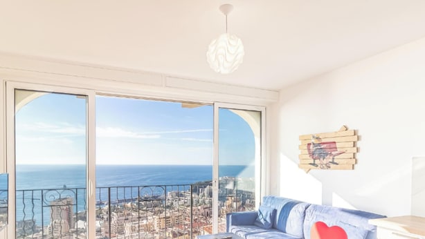 A room with a large window offering a city view, a blue sofa, and a wooden wall decoration. - Amazing Sea View T2- Near Monaco (Beausoleil)
