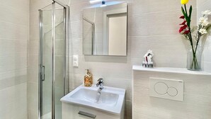 Uma casa de banho moderna com cabine de duche em vidro, lavatório branco com espelho em cima e um vaso de flores na parede.