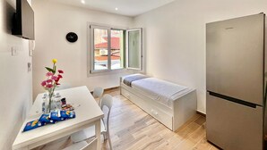 Quarto compacto com uma cama, uma mesinha com um vaso e petiscos, uma TV e um frigorífico.