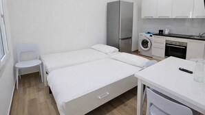 Quarto pequeno e limpo com uma cama, uma cadeira, uma mesa, uma máquina de lavar e um frigorífico.
