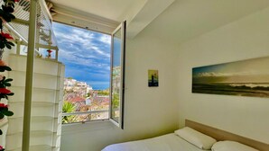 A bedroom with a bed, a window with a view of buildings, a painting on the wall, and a white shelf with decorative items. - T2 Splendid View Near Monaco (Beausoleil)