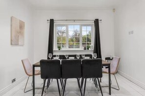 A dining area with a table set for six, black chairs, a window with curtains, and a view of greenery outside. - Perfect Booking 4 Bed Home in Cheltenham (Cheltenham)