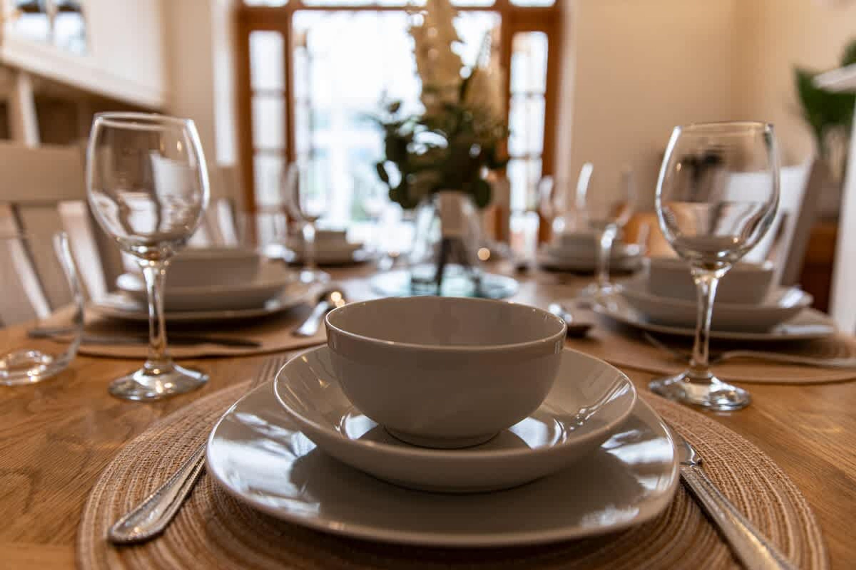 Mesa de jantar posta com pratos, tigelas e copos de vinho brancos, mesa de madeira e um jogo americano tecido.