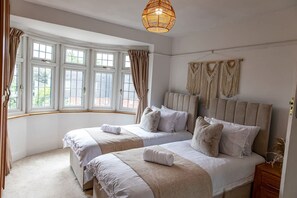 A bedroom with two beds, a large window with curtains, a woven light fixture, and macramé wall hangings.