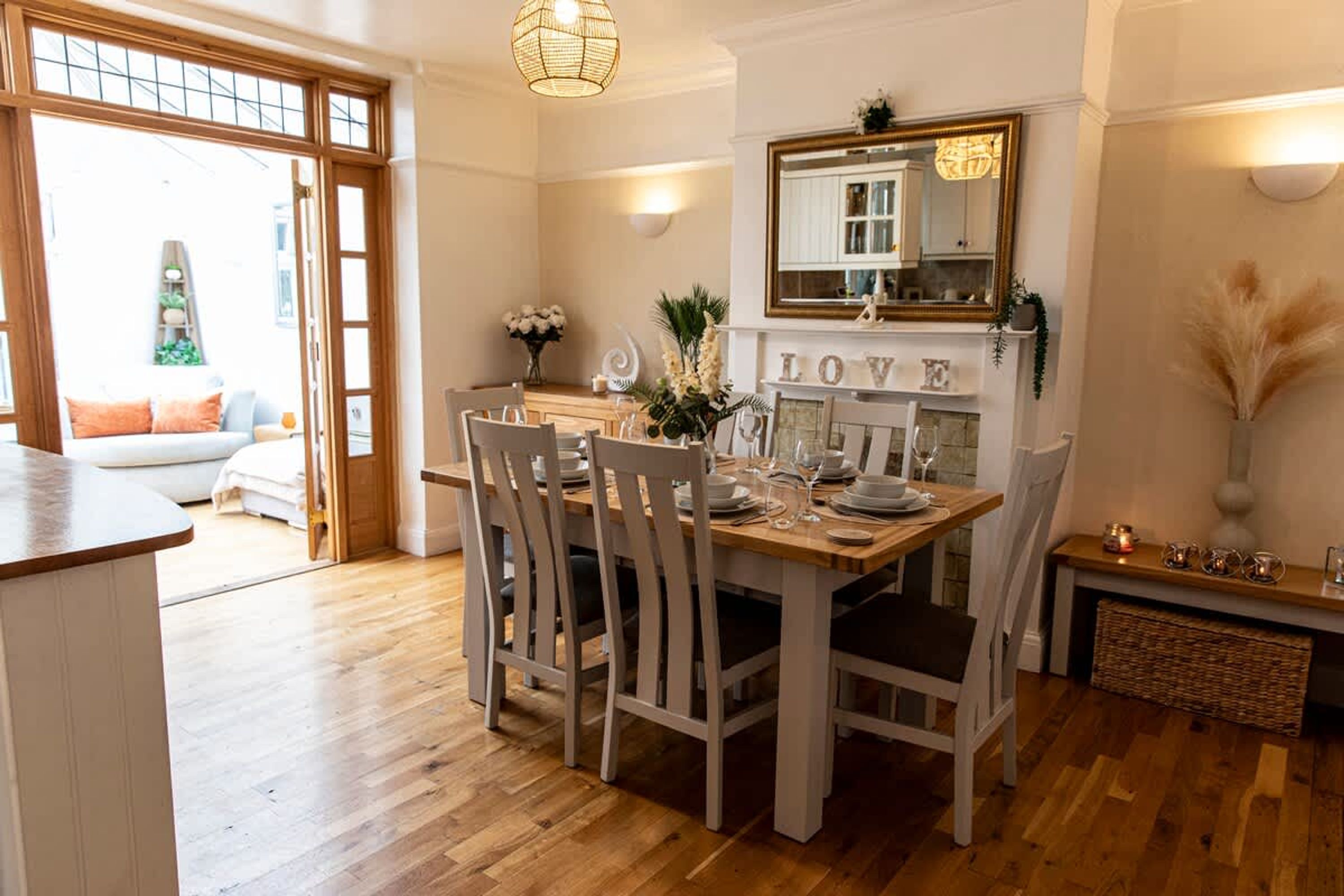 A dining area with a wooden table set for four, white chairs, a mirror above the fireplace, and a 'LOVE' sign on the mantel.