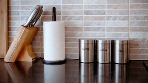 Un plan de travail de cuisine avec un porte-essuie-tout, un bloc à couteaux et des récipients en acier inoxydable étiquetés « CAFÉ », « SUCRE » et « THÉ ».