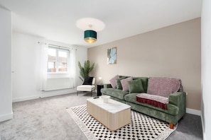 A living room with a green sofa, a white coffee table, a patterned rug, a potted plant, and a ceiling light fixture. - Perfect Booking 3 Bed With Driveway Parking (Gloucester)