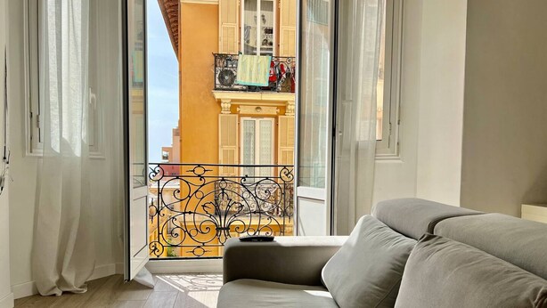 A modern living room with a sofa, a glass door leading to a balcony, and a view of a building with shutters. - Stylish 3-bedroom Gem Steps From Monaco Spacious (Beausoleil)