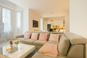 Ein modernes Wohnzimmer mit einem beigen Sofa, einem Glastisch und einer Küche im Hintergrund.