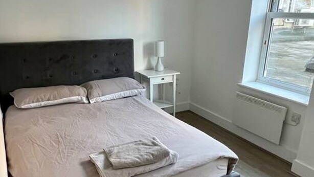 A neatly made bed in a room with a window, a bedside table with a lamp, and a radiator. - Lovely 2-bed Flat With Great Transport Links (London)