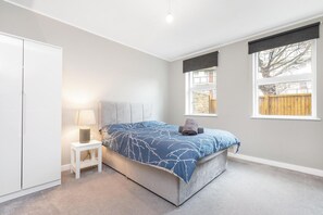 A bedroom with a bed, a nightstand, a lamp, and a window with a view of a fence. - Spacious 2-bed Flat With Outdoor Patio (London)