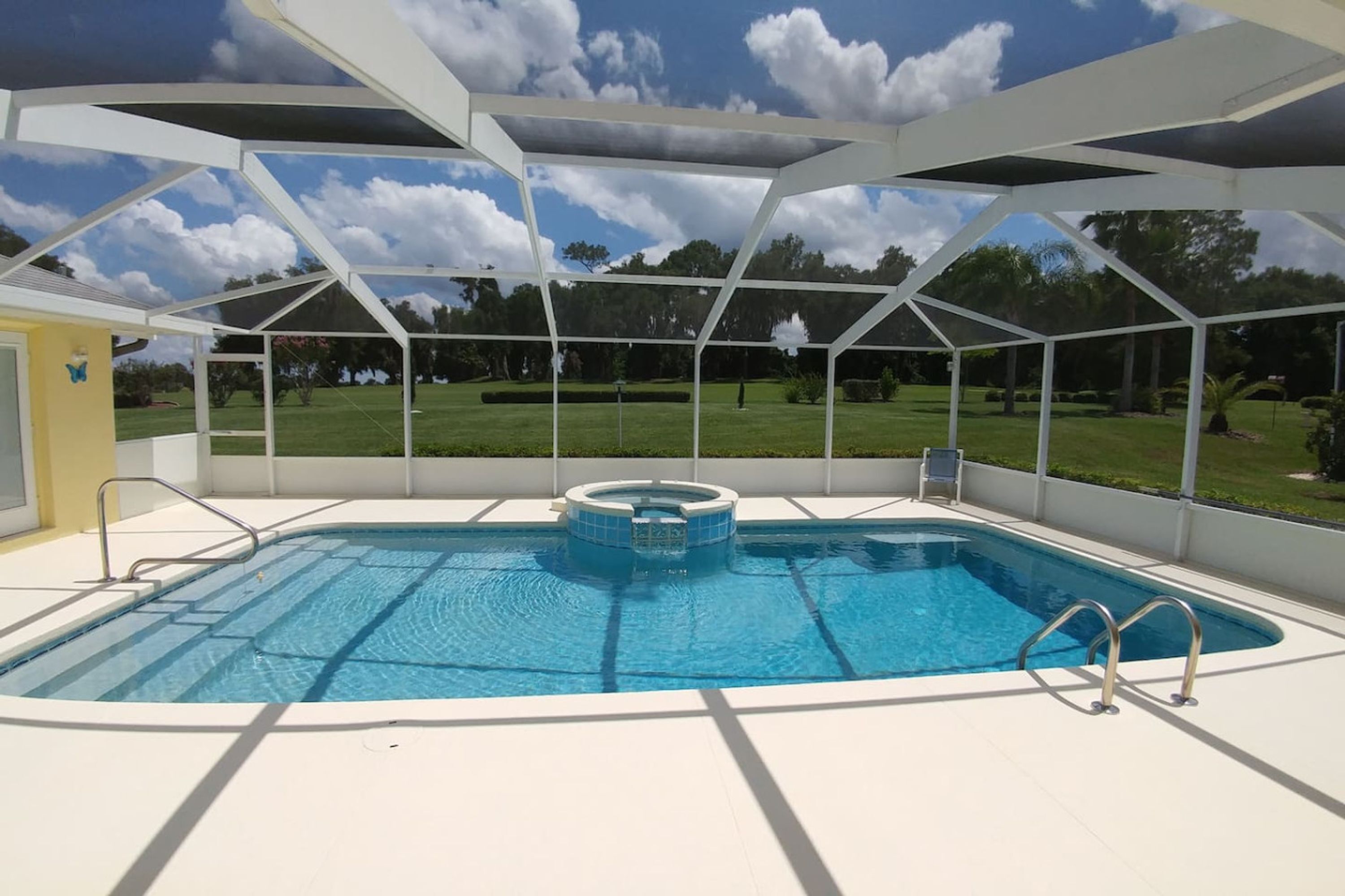 Une piscine dotée d’une structure servant de parasol, entourée d’une terrasse blanche et offrant une vue sur une zone verdoyante.