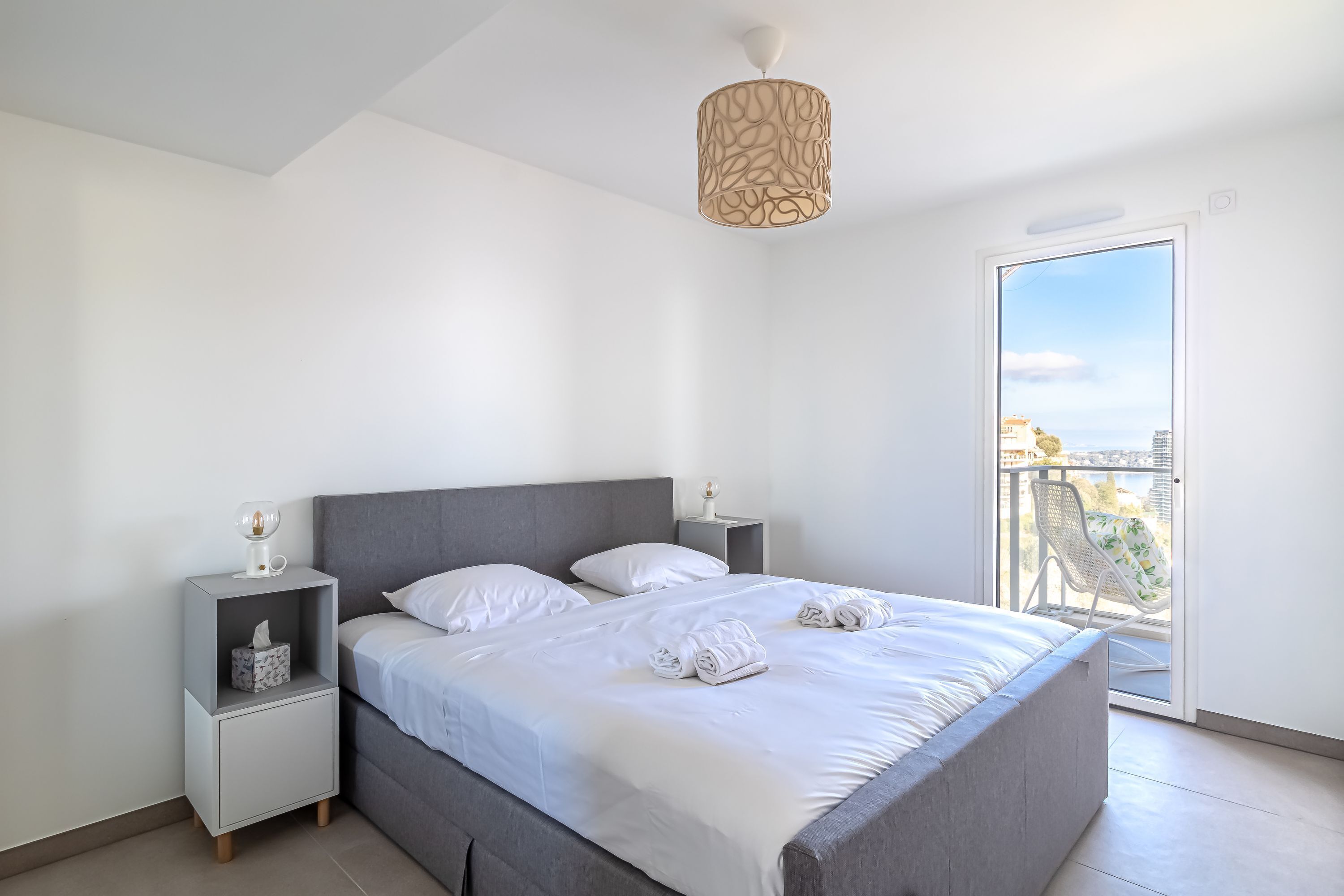 Una camera da letto moderna con un letto grigio, lenzuola bianche, un comodino con una lampada e vista sulla città tramite una porta finestra.