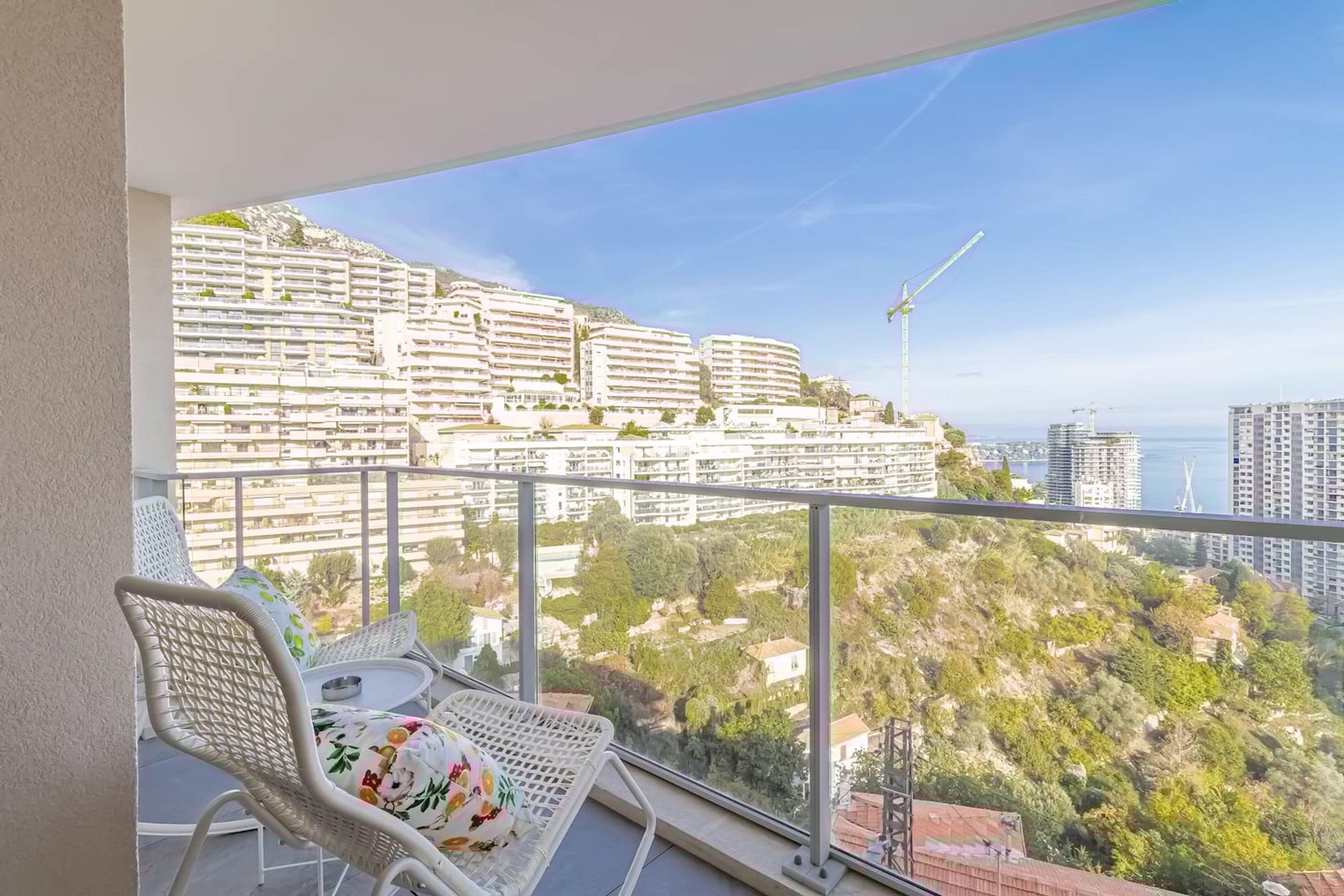 A balcony with a white chair and a table, overlooking a cityscape with buildings and a wind turbine.