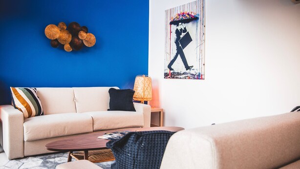 Een woonkamer met een blauwe accentmuur, een beige bank, een houten salontafel en een kamerkleed met een patroon.