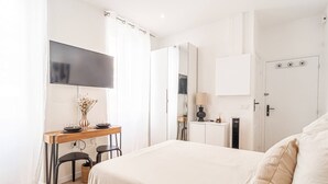A bedroom with a bed, a desk with chairs, a TV, a mirror, a door with a lock, and a small cabinet. - Nice Studio Close to Monaco (Beausoleil)