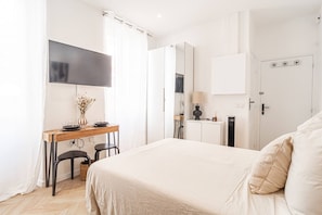 A bedroom with a bed, a desk with chairs, a TV, a mirror, a door with a lock, and a small cabinet. - Nice Studio Close to Monaco (Beausoleil)