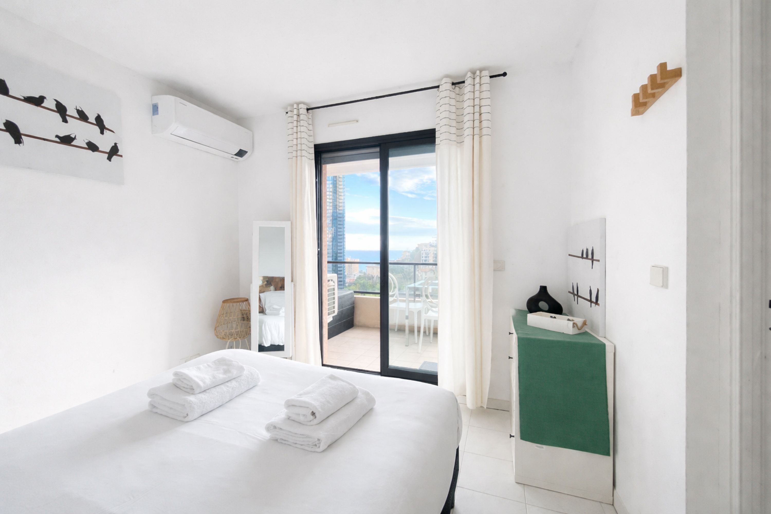 A hotel room with a bed, a green vanity, a balcony with a view, and a wall-mounted air conditioner.
