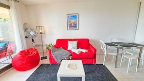 Een moderne woonkamer met een rode bank, een witte salontafel, een eettafel met stoelen, een ventilator en een poster aan de muur.