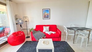 A modern living room with a red sofa, a white coffee table, a dining table with chairs, a standing fan, and a poster on the wall. - 1BR Sea Monaco View Terrace (Beausoleil)