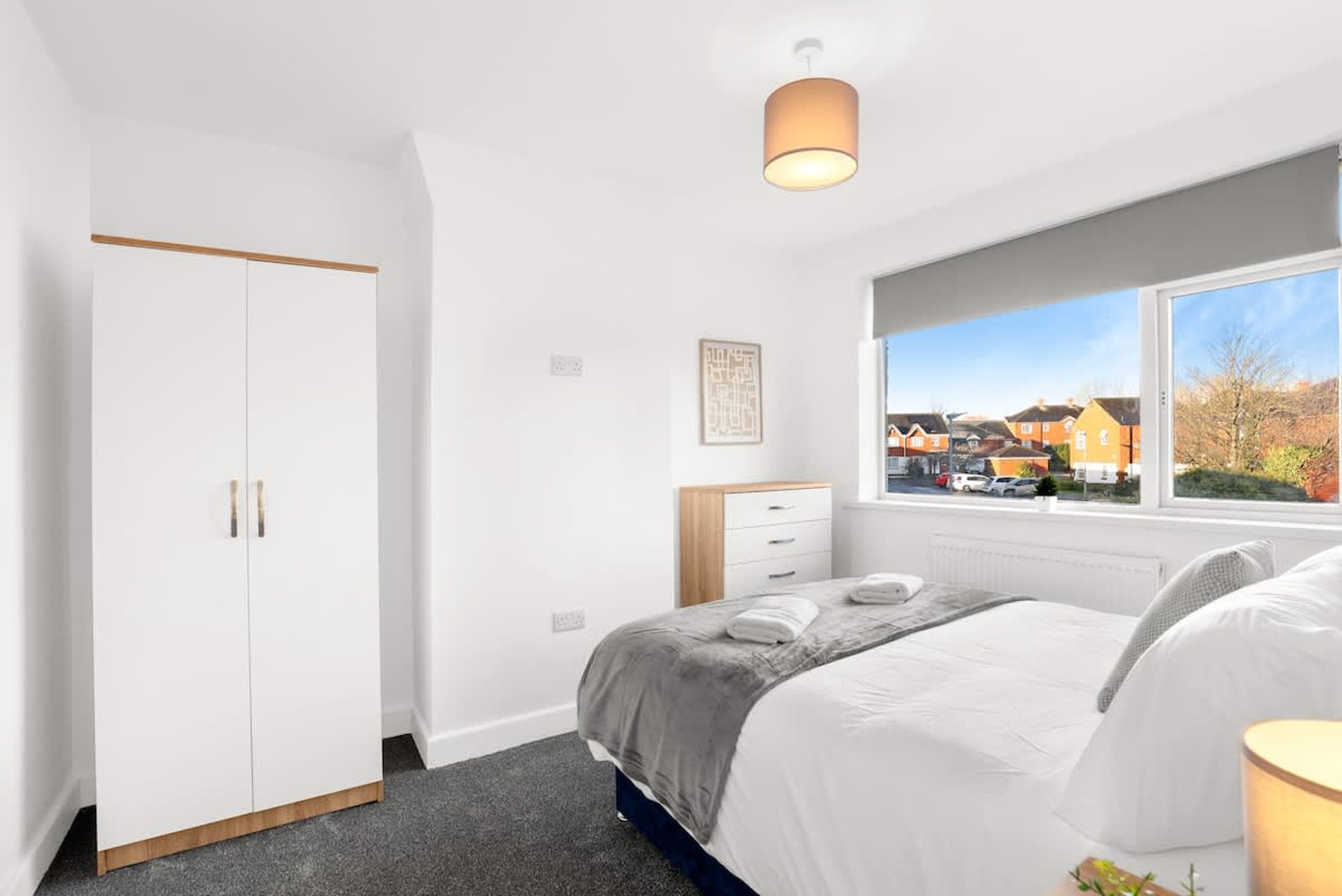 A bedroom with a bed, a wardrobe, a window with a view of houses, and a ceiling light.