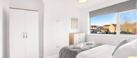 A bedroom with a bed, a wardrobe, a window with a view of houses, and a ceiling light.