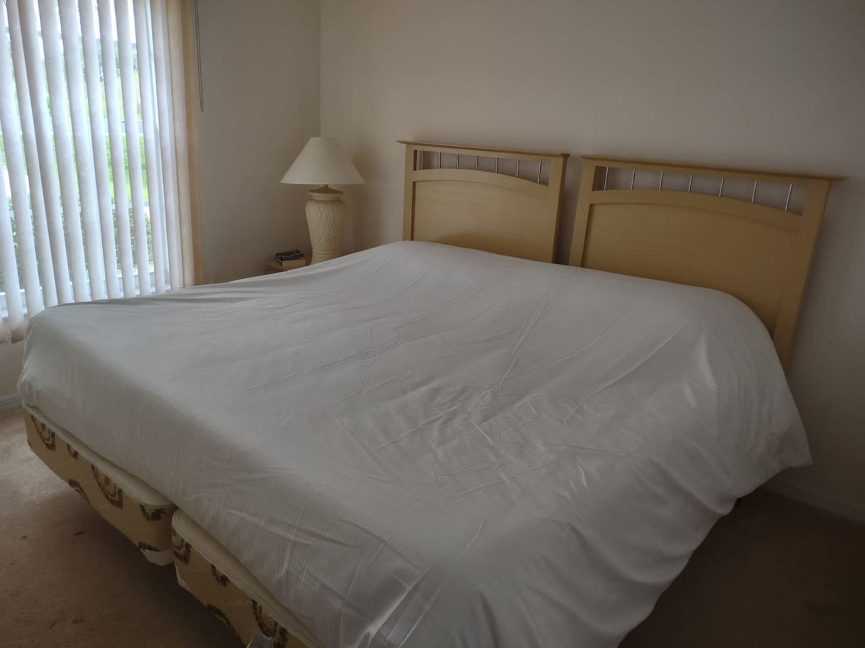 Een bed met een witte spreidek, een nachtkastje met een lamp en een raam met blinds.