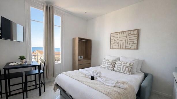 A bedroom with a bed, a desk with a chair, a TV, a wardrobe, and a view of the sea. - Bright Studio Flat in Cap d Ail - Near Monaco (Cap d'Ail)