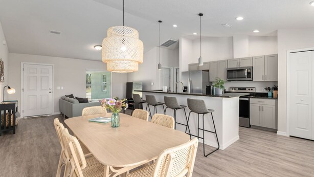 A modern kitchen with white cabinets, a central island, and a dining area with a wooden table and wicker chairs. - Villa Verde Cozy Home In New Community (Inverness)