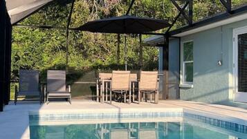 A swimming pool with a covered patio area, outdoor chairs, and a view of trees.