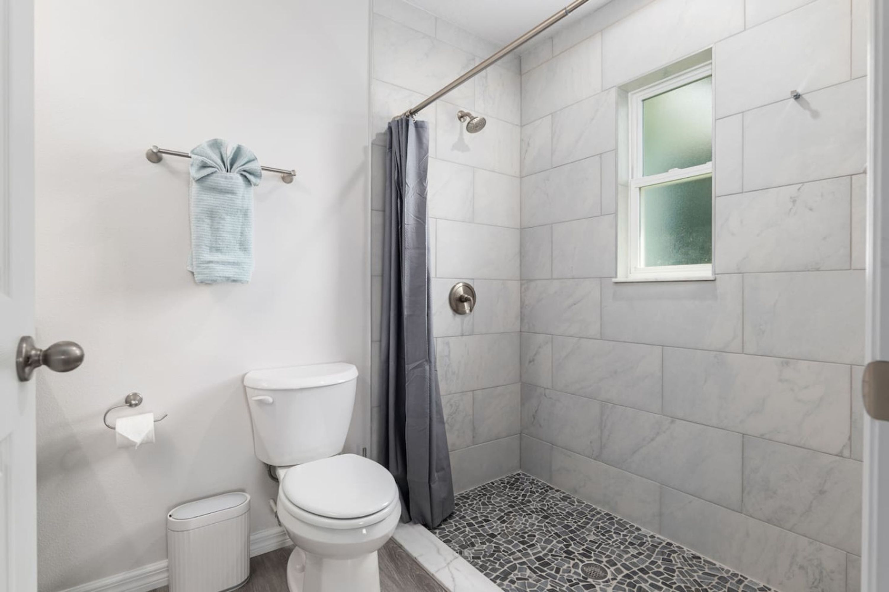 A bathroom with a toilet, a shower with a curtain, a window, and a towel rack.