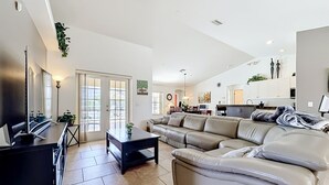 A spacious living area with a large sofa, a coffee table, and a TV. There is a kitchen in the background with cabinets and a dining area. - Villa Havelaar Family Friendly and Awesome Pool (Inverness)