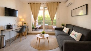 Ein Wohnzimmer mit einer Couch, einem Couchtisch, einem Fernseher, einem Schreibtisch und einem Balkon mit Vorhängen.