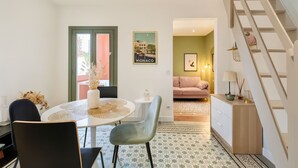 Un coin repas avec une table ronde, des chaises et un vase rempli de fleurs séchées. Un tableau encadré orne le mur, et un escalier mène à une autre pièce.