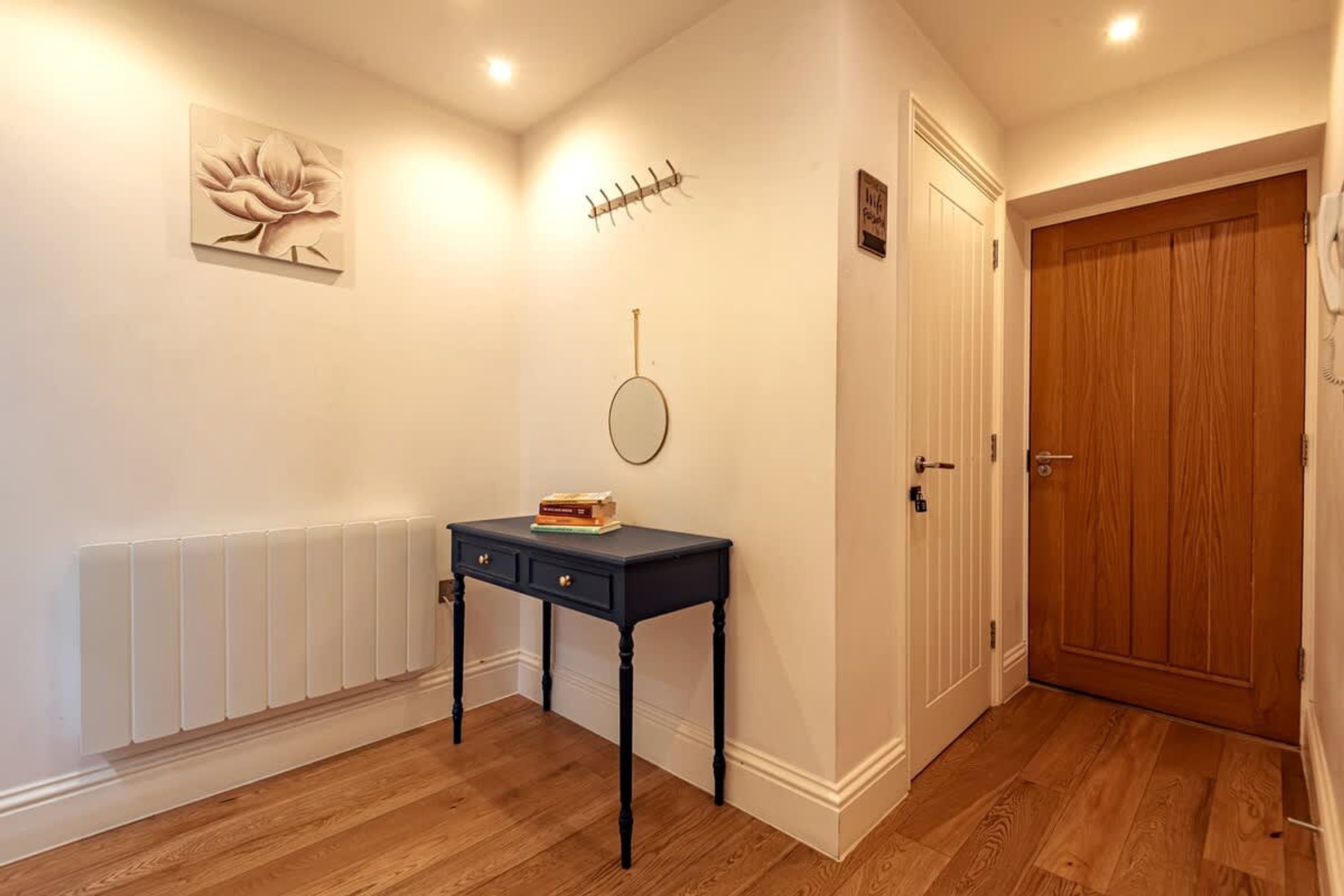 A hallway with a radiator, a wooden door, a wall-mounted coat rack, a round mirror, and a small table with books.