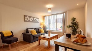 Un salon avec un canapé, une table basse, une table à manger avec des croissants, une lampe et une fenêtre avec des rideaux.