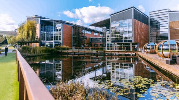 Un bâtiment moderne doté d’une façade en verre, d’un plan d’eau réfléchissant et d’une terrasse en bois.