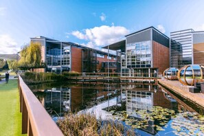 Een modern gebouw met een glazen gevel, een reflecterend waterlichaam en een houten terras.