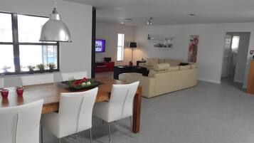 A modern living room with a sofa, a dining area with chairs and a table, and a television in the background.