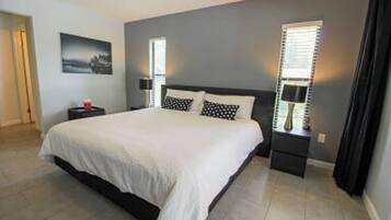 A bedroom with a bed, two bedside tables, a nightstand, a window with blinds, and a wall-mounted lamp.