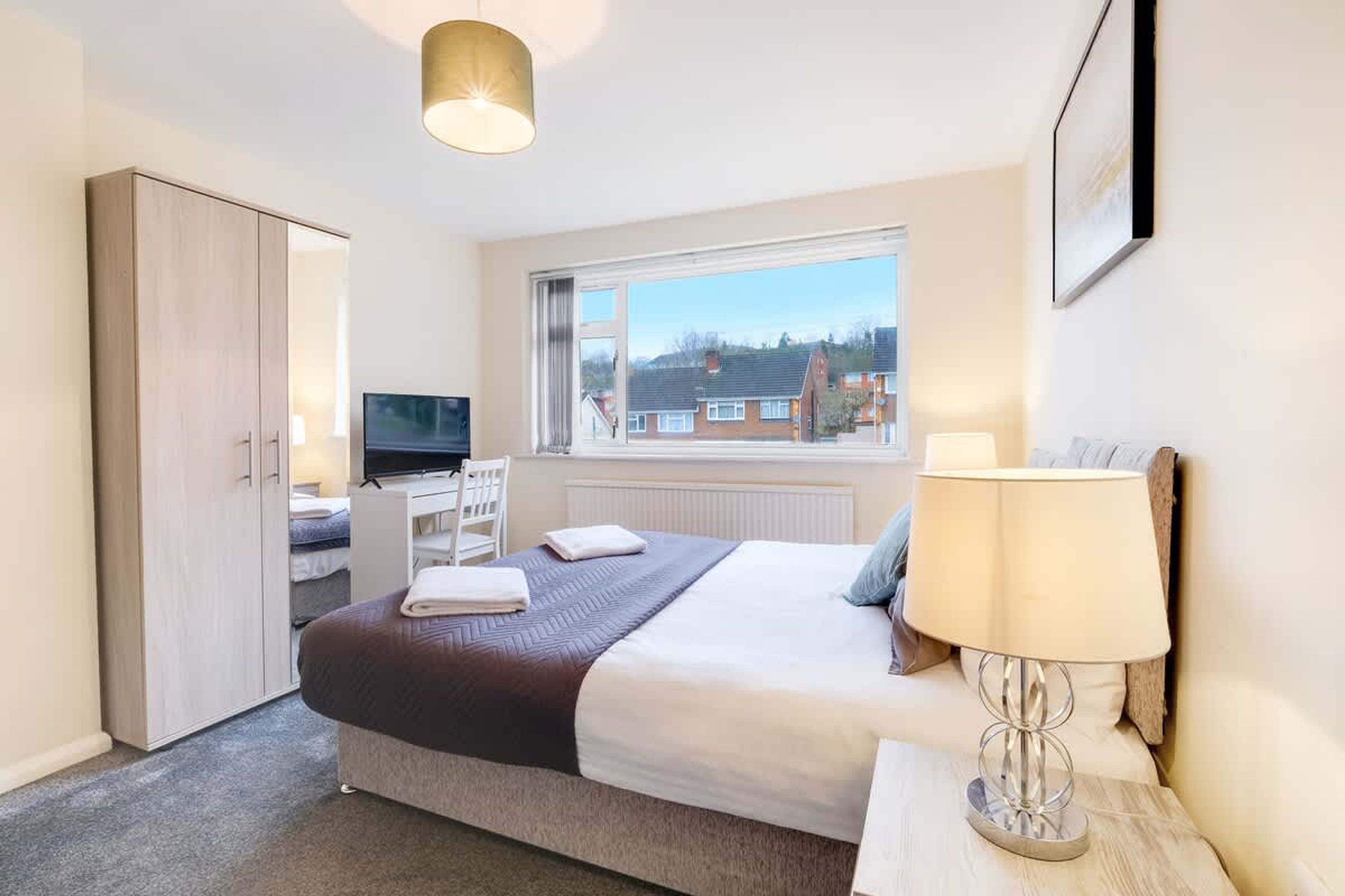 A hotel room with a bed, a desk, a TV, a window with a view, and a lamp.