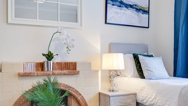 A bedroom with a bed, a nightstand, a lamp, a wall-mounted shelf with a plant, and a framed picture. - High Wycombe Home Free Parking Free Wifi Garden (High Wycombe)