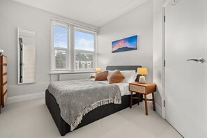 Ein Schlafzimmer mit Bett, Nachttischen, einem Fenster, einer Tür und einem Wandbild.
