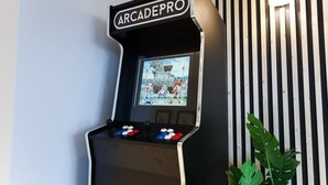 Una máquina de videojuegos con la etiqueta 'ARCADEPRO', una pantalla mostrando un juego, controles de joystick y una planta en maceta a su lado.