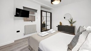 Una camera da letto moderna con un letto, una TV a parete, un termosifone, una porta scorrevole in vetro, una cassettiera scura e uno specchio rotondo.