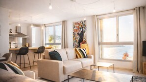 Een moderne woonkamer met een bank, salontafel en barkrukken. Een keuken is zichtbaar met een fornuis en aanrecht. Grote ramen bieden uitzicht op zee.