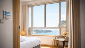 Een slaapkamer met een bed, een klein rond tafeltje, een stoel, een lamp en uitzicht op zee.