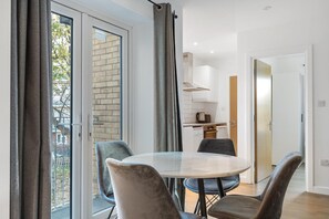 A modern apartment with a dining area featuring a round table and four chairs, a kitchen with white cabinets, and a sliding glass door leading to a balcony. - Modern Flat in Prime Location (London)