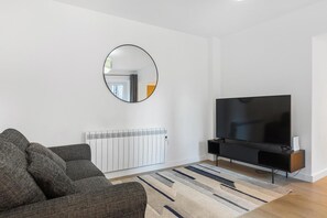 Een moderne woonkamer met een grote flatscreen-tv, een grijze bank, een ronde spiegel en een radiator.